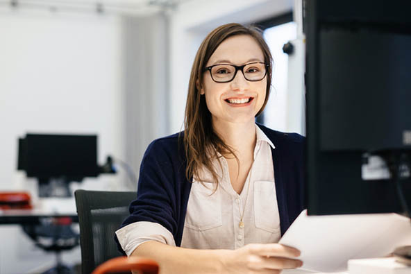 A young woman sits at her office desk smiling into the camera. She has light brown, shoulder length hair, wears glasses and is dressed in a pale off-white shirt and dark blue cardigan. She's holding papers. A desk and computer are behind her, out of focus