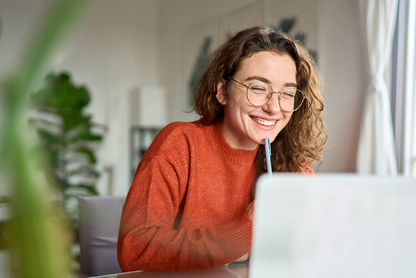A smiling woman wearing glasses and an orange sweater, works at her computer. She holds her pen up against her chin. The room is modern with a tall lamp and plants in the background