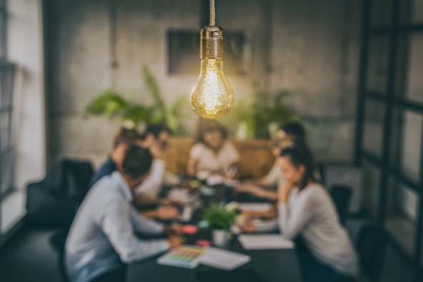 Behind a glowing lightbulb, a group of business people are gathered around a table discussing a new idea.